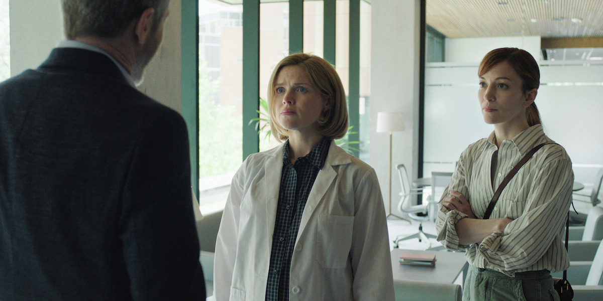 Two women, one in a lab coat and one in casual attire, stand in a modern office or lab with large windows, talking to a man whose back is to the camera.