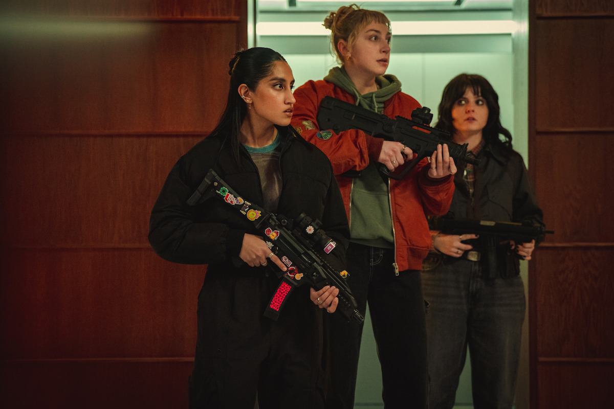 Three women stand alert in a modern building hallway, holding toy-like guns and wearing serious expressions, suggesting a tense or adventurous scenario.