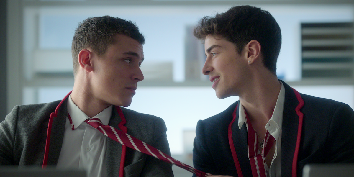 Two young men in school uniforms sit indoors, one gently holds the other's striped tie while they exchange a meaningful look, suggesting a close or flirtatious interaction, with shelves and books in the background.