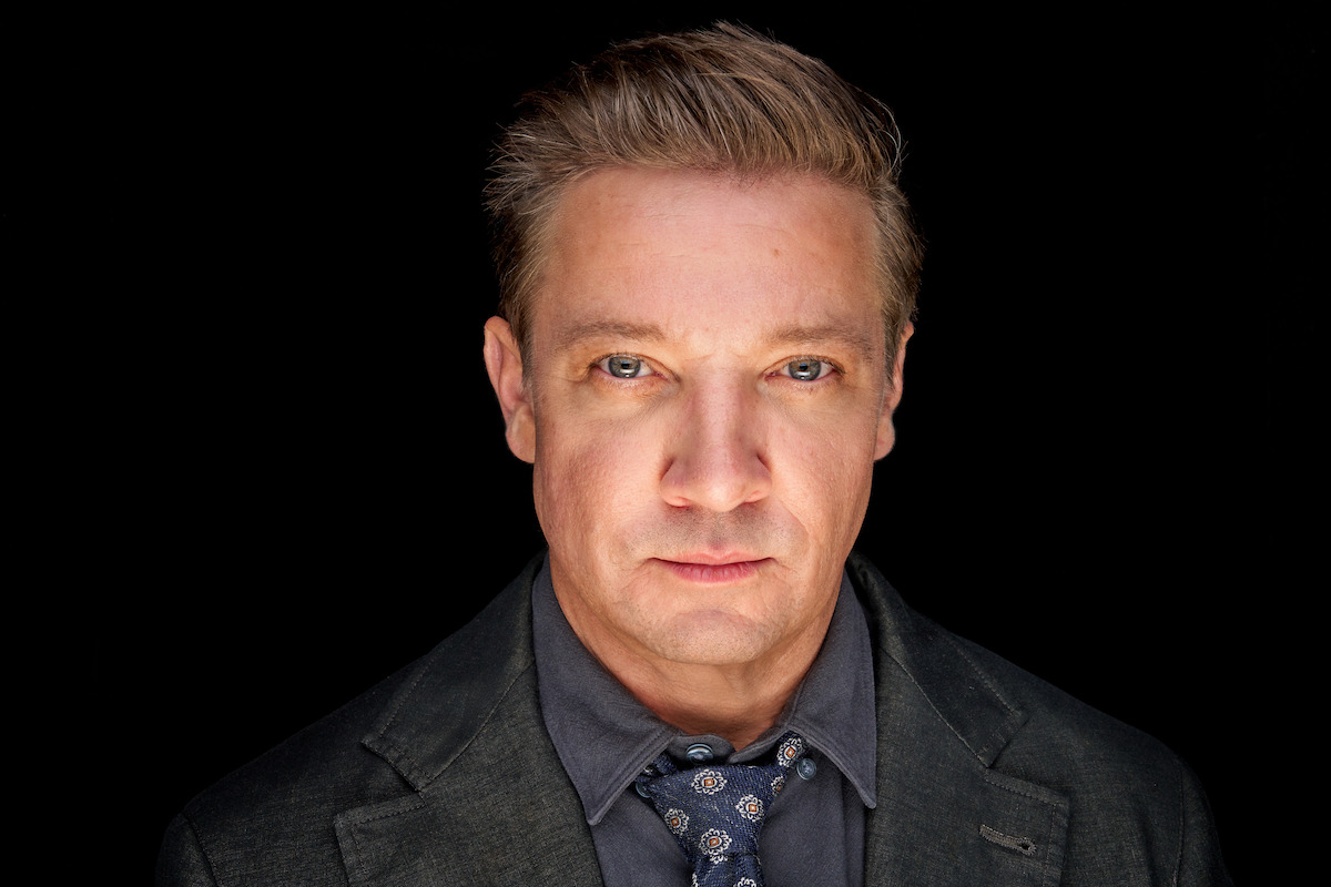A serious-looking man in a suit and patterned tie stands against a black background, illuminated by direct lighting, creating a dramatic and intense mood in a studio portrait setting.