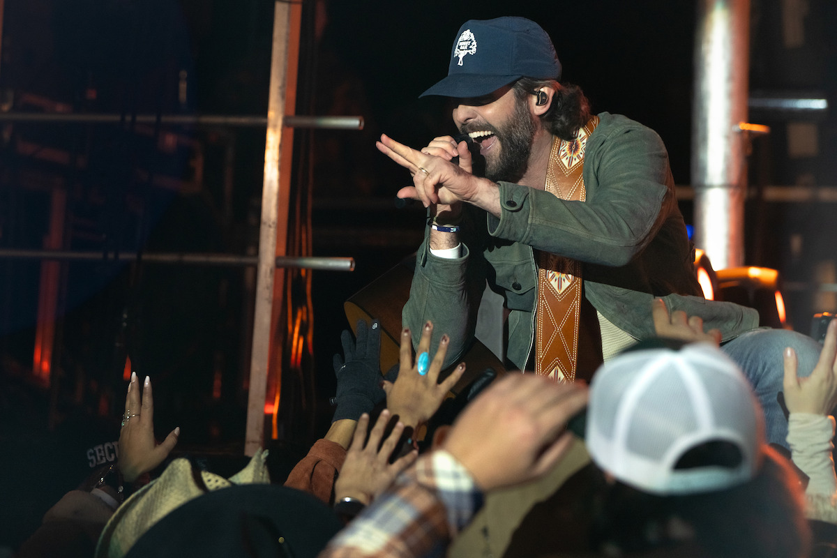 Male singer wearing a cap and casual jacket performs energetically on stage, reaching out to enthusiastic fans in the front row, with concert lighting and metal scaffolding visible in the background.