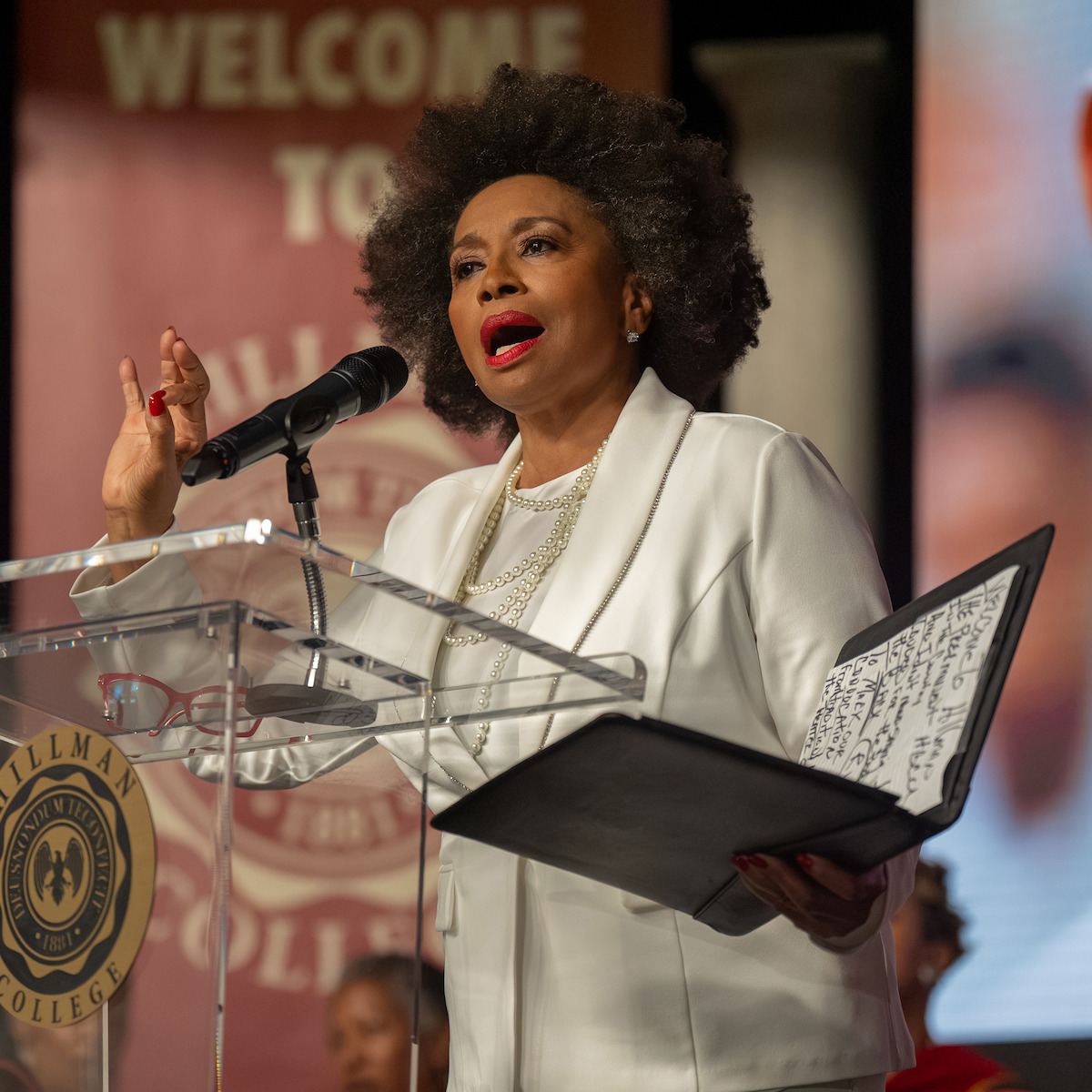 A woman in a white suit speaks passionately at a podium with a microphone, holding a folder with notes. The event is at a college, indicated by banners and the college emblem on the podium, with an audience in the background.