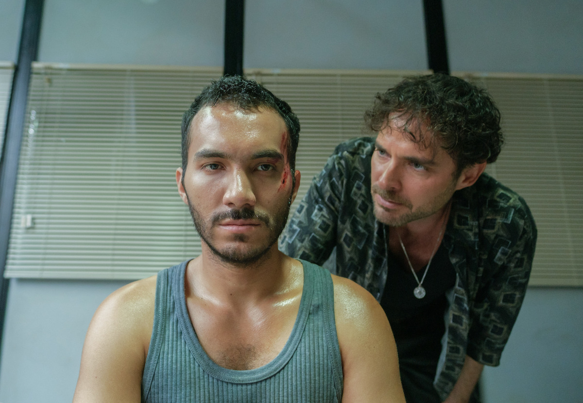 A man wearing a green tank top with a bloodied face stands in front of Manolo Cardona as Danger in a police interrogation room in an image from the series ‘Medusa.’