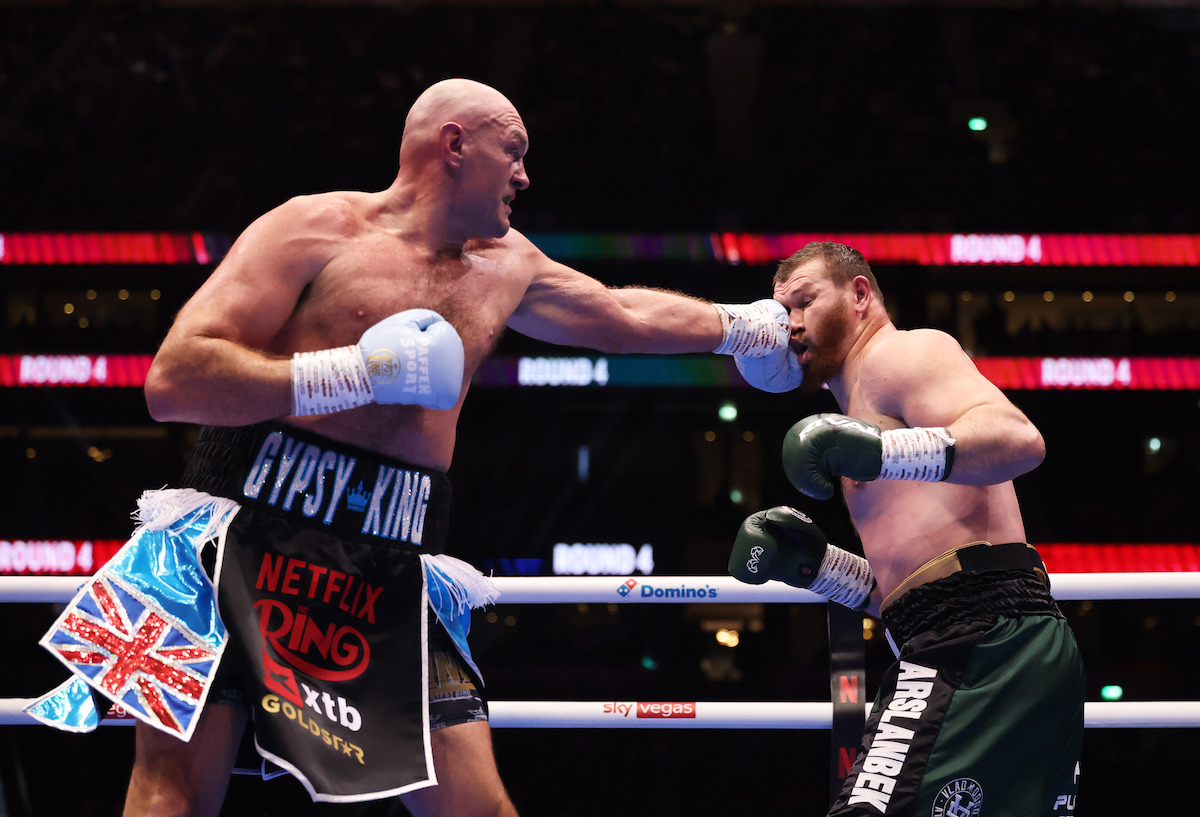 Two heavyweight boxers fight in a brightly lit boxing ring, one landing a punch to the other's face. They wear gloves, shorts, and are surrounded by an audience in a large, darkened arena.