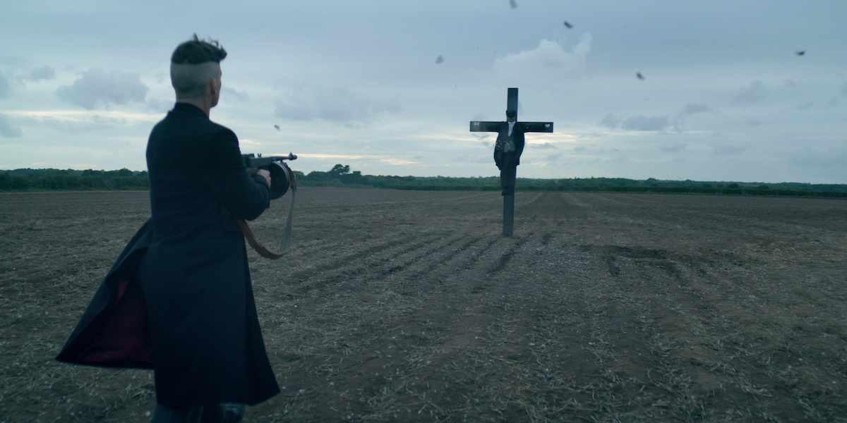 A man with closely cropped hair fires a gun at a scarecrow in an otherwise empty field.