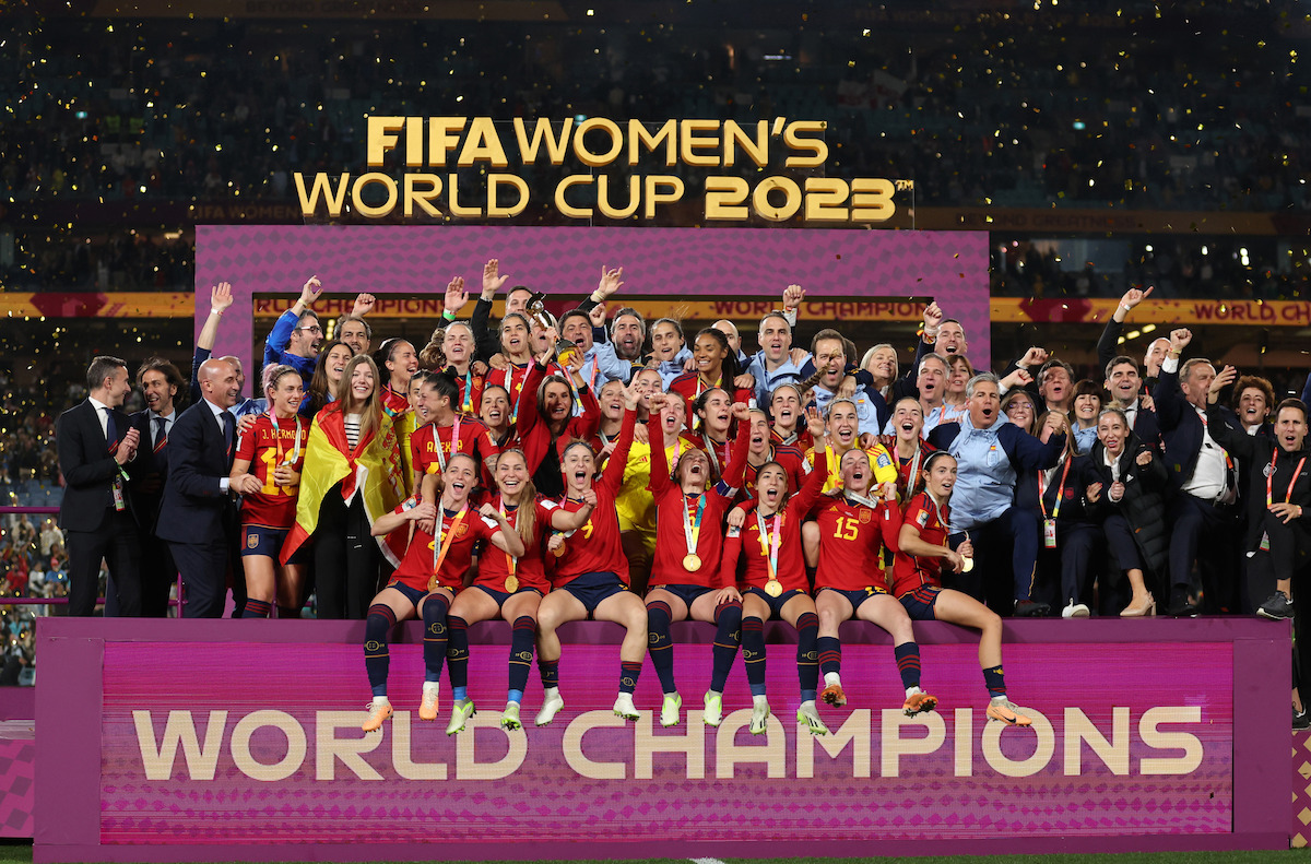 Soccer team celebrating victory on stage with confetti, FIFA Women's World Cup 2023 sign above, and "World Champions" banner in stadium.