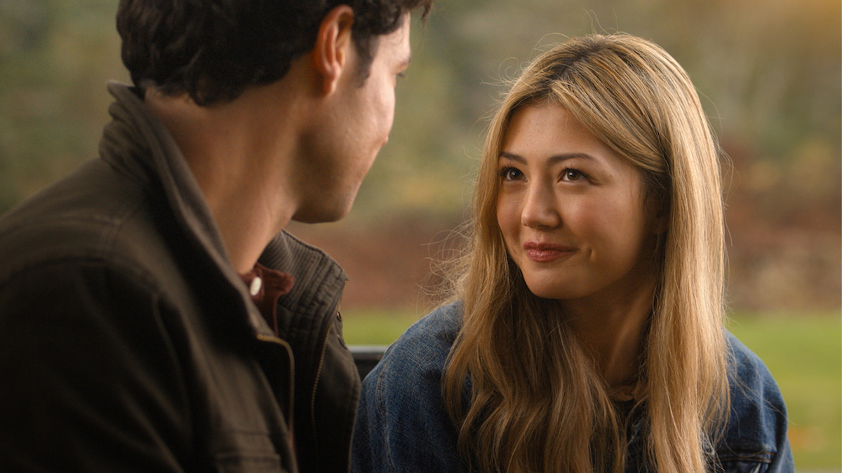 A young woman with long blonde hair smiles warmly at a man while sitting outdoors in a park-like setting with blurred greenery in the background, suggesting a friendly or romantic conversation.