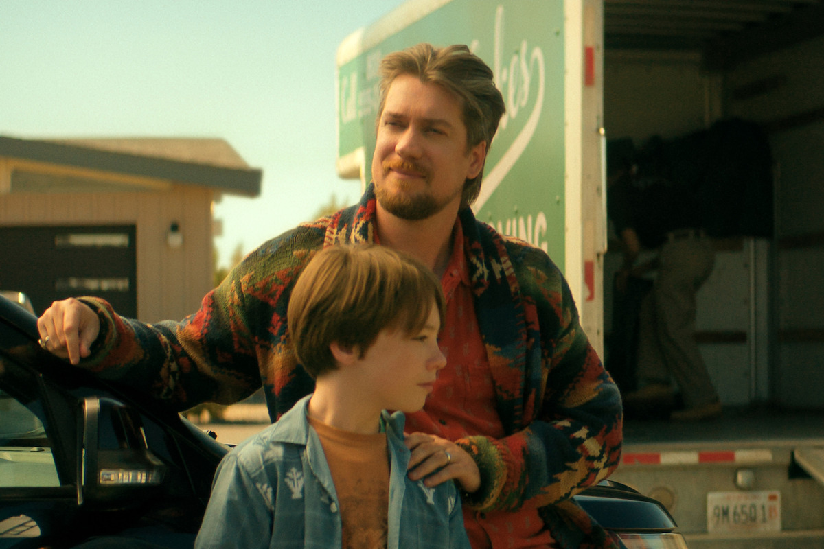 Rafeal Casal as Neil in a colorful sweater stands beside young boy near black car, with moving truck and house in the background on a sunny day, suggesting a moving or arrival scene in a suburban neighborhood.