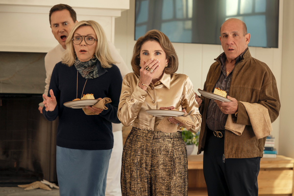 Four adults indoors, holding dessert plates, look shocked or surprised, standing near a fireplace and TV in a modern living room with natural light.