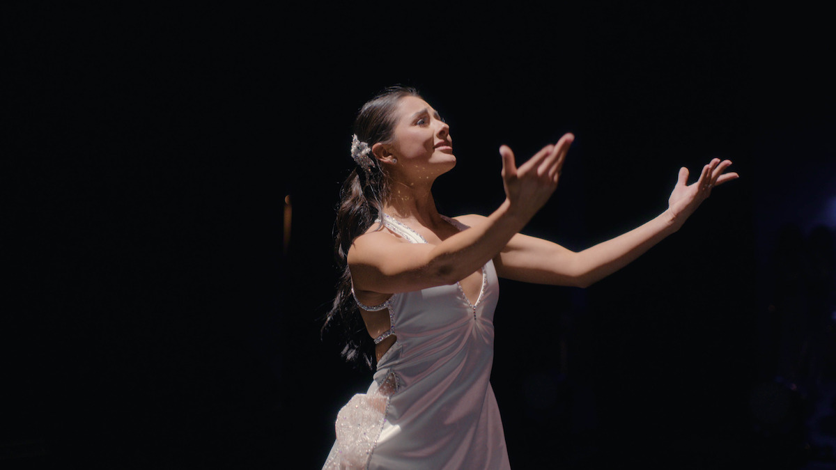 A woman in a white dress stands on a dark stage under a spotlight, reaching out with expressive hands, appearing to sing or perform dramatically.