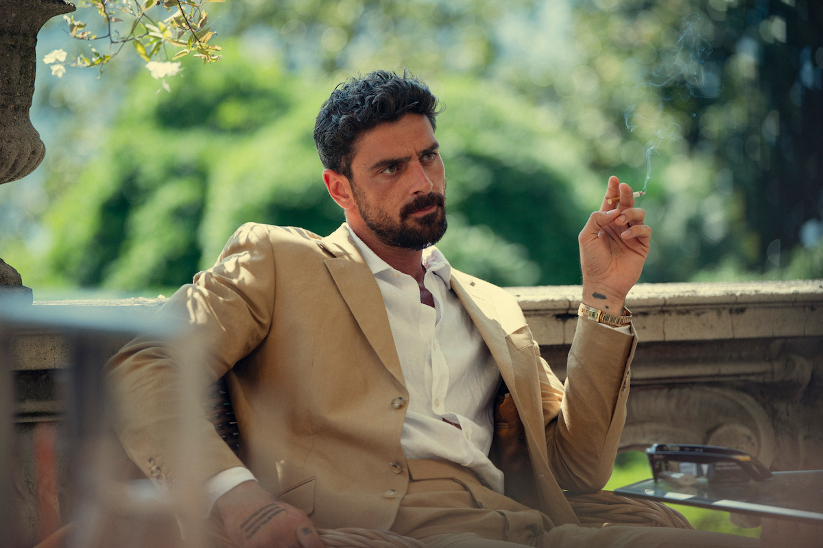 Man in a tan suit sitting outdoors on a stone terrace, surrounded by greenery, holding a cigarette with a relaxed and contemplative expression.