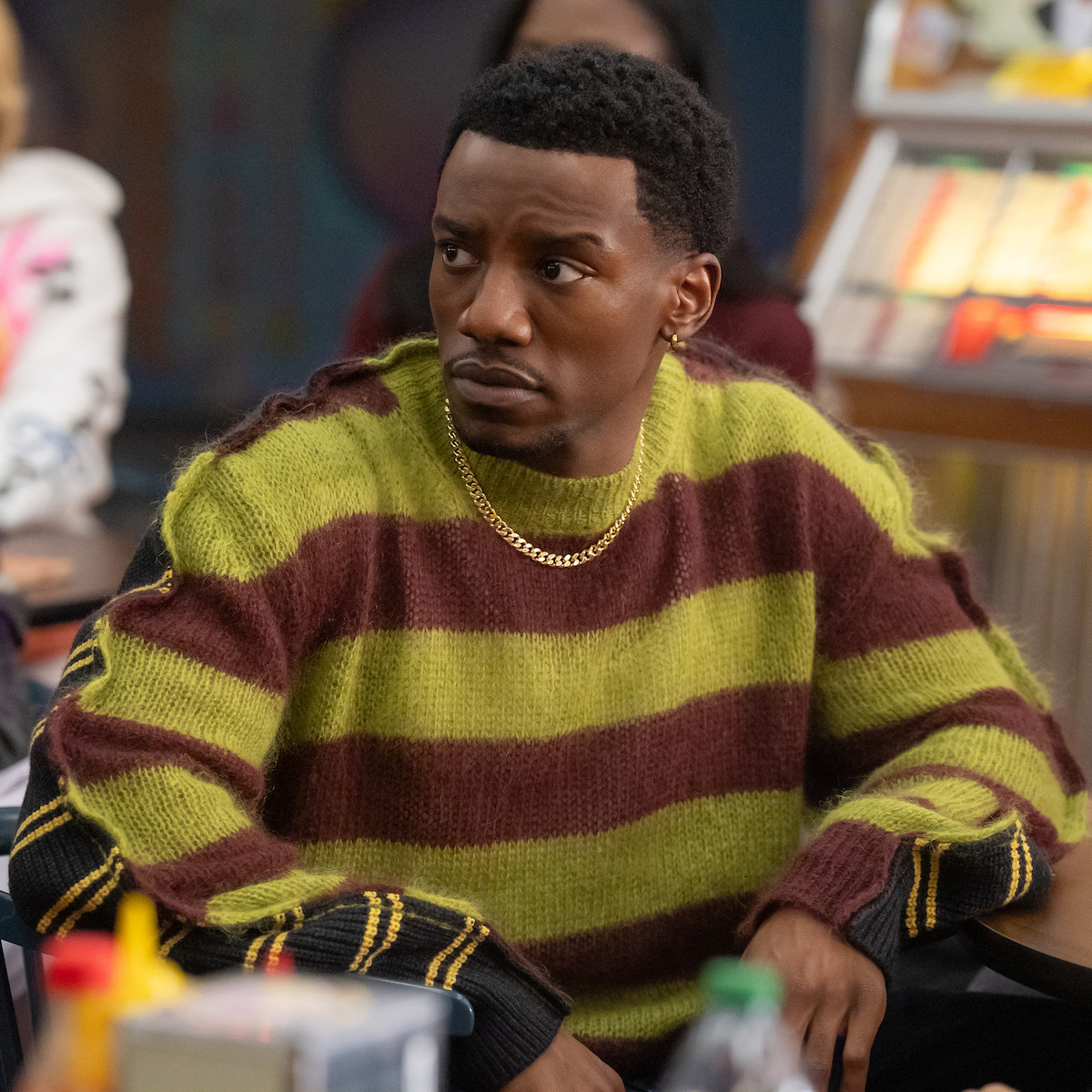 Man in a green and brown striped sweater sits at a table in a classroom setting, looking to the side with a serious expression. Other people and classroom items are visible in the background.