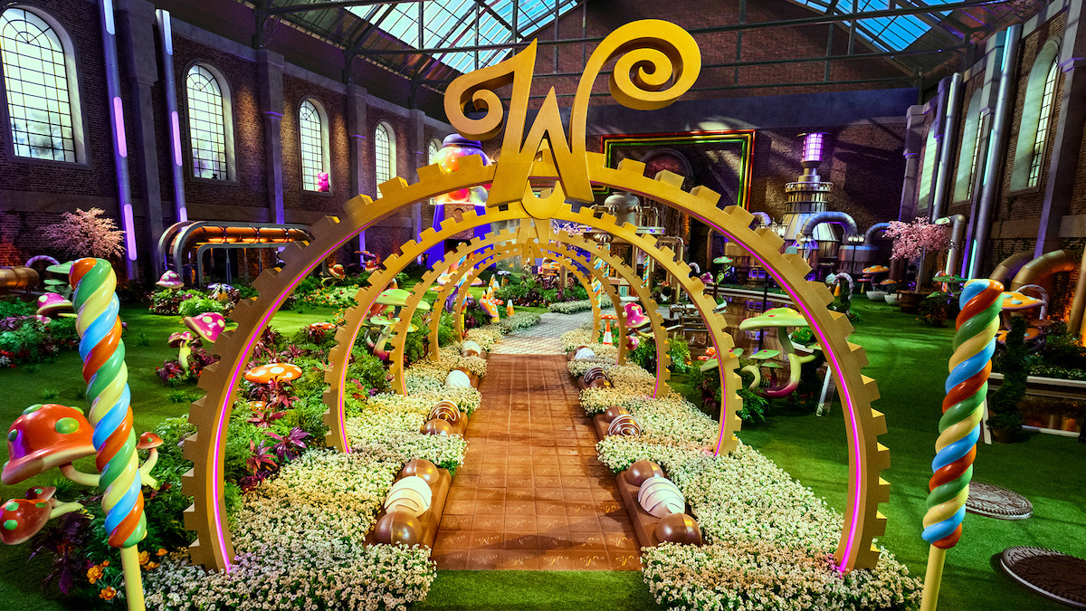 A whimsical candy-themed garden inside a large factory with colorful sweets, oversized mushrooms, lollipops, golden arches, flowers, and a large "W" centerpiece under a glass roof, creating a magical, playful atmosphere.