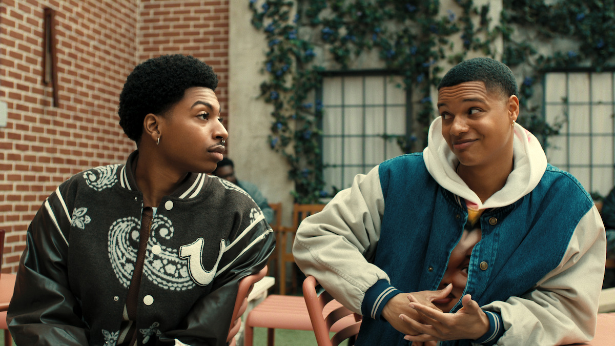 Two young men sit outside on pink chairs against a brick wall and windows with green vines, looking at each other and smiling, wearing casual jackets in a relaxed courtyard setting.
