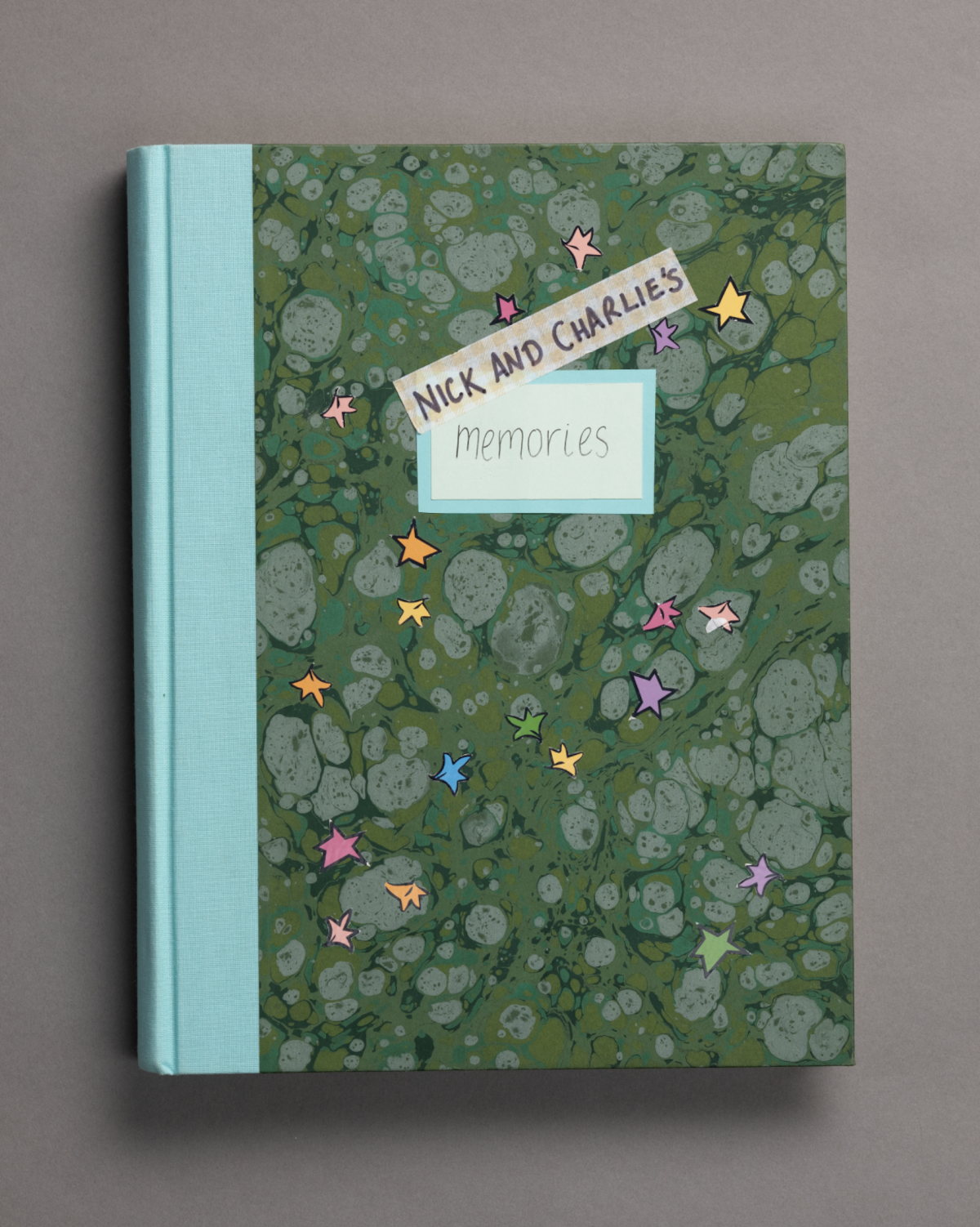 A scrapbook with a marbled green cover, light blue spine, and label reading "Nick and Charlie's memories," decorated with colorful star stickers, resting on a gray surface.