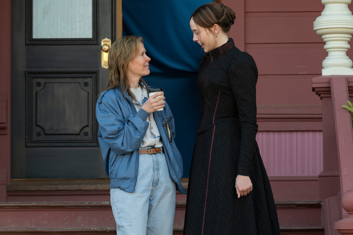 Two women talking on a porch, one in a blue jacket with jeans holding a coffee cup, the other in a long black dress, in front of a maroon house with wooden steps and columns.