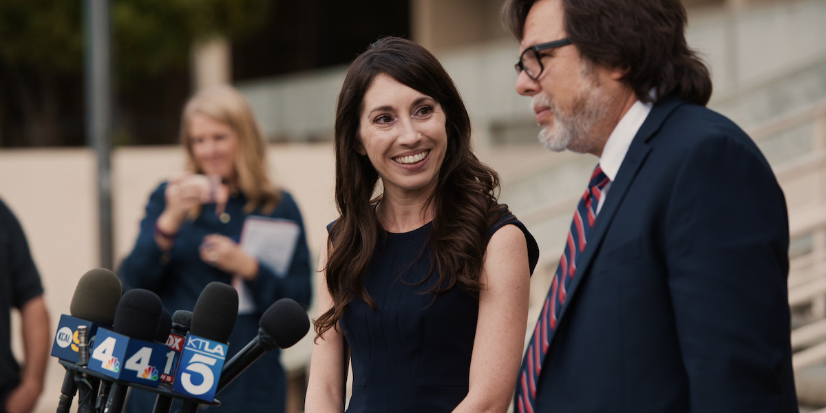 A woman and a man stand in front of microphones labeled with news station logos, smiling and talking outside on a staircase. Reporters and news crew are visible in the background, creating a professional and public setting.
