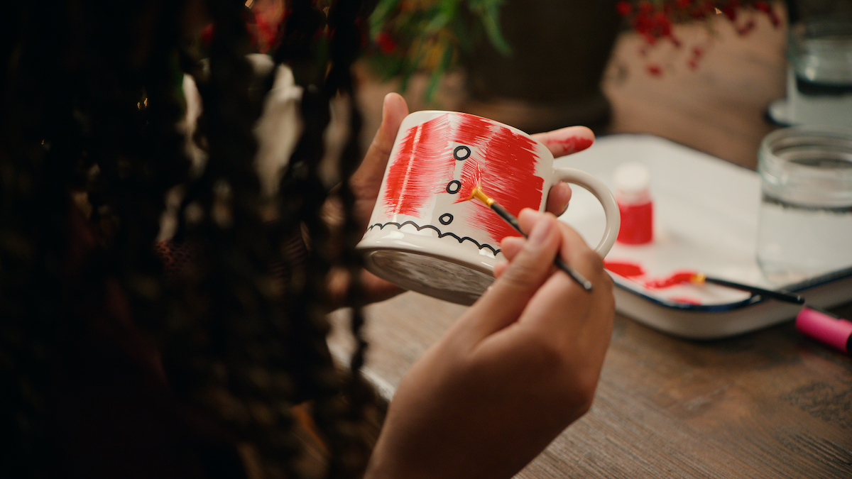 Person painting a festive red mug with black buttons and white scalloped edge detail at a cozy table, art supplies and water nearby, creating a warm and creative atmosphere.