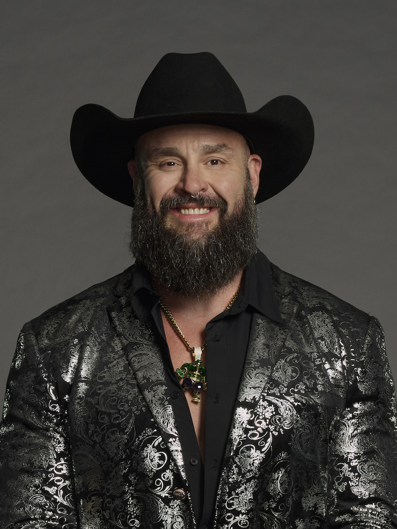 Man with a beard wearing a black cowboy hat, black patterned blazer, and necklace, standing in front of a plain, dark background, smiling at the camera in a studio setting.