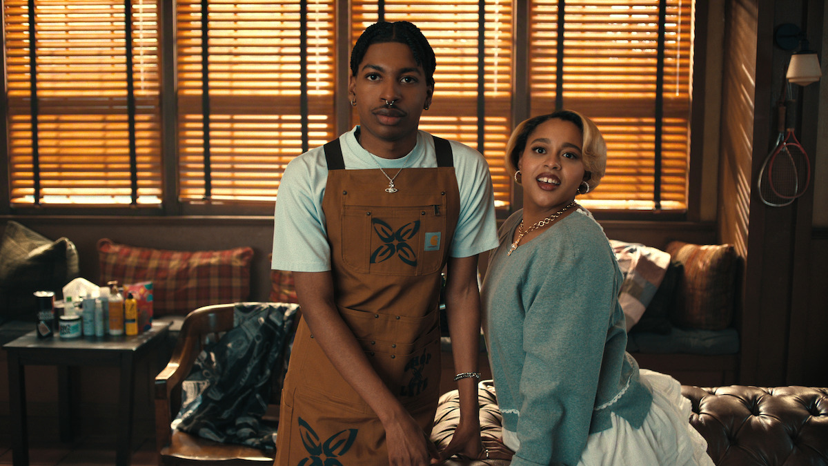 Two people stand together in a warmly lit room with wooden blinds. One wears a brown apron and white shirt, the other a gray top and white skirt. The setting is cozy, with a couch, table, and scattered items in the background.