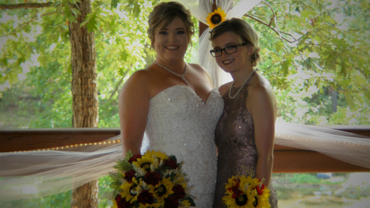 Two women dressed formally, one in a white wedding gown and the other in a sparkly dress, smiling and holding sunflower bouquets, standing by a wooden railing with greenery and soft drapery in the background.