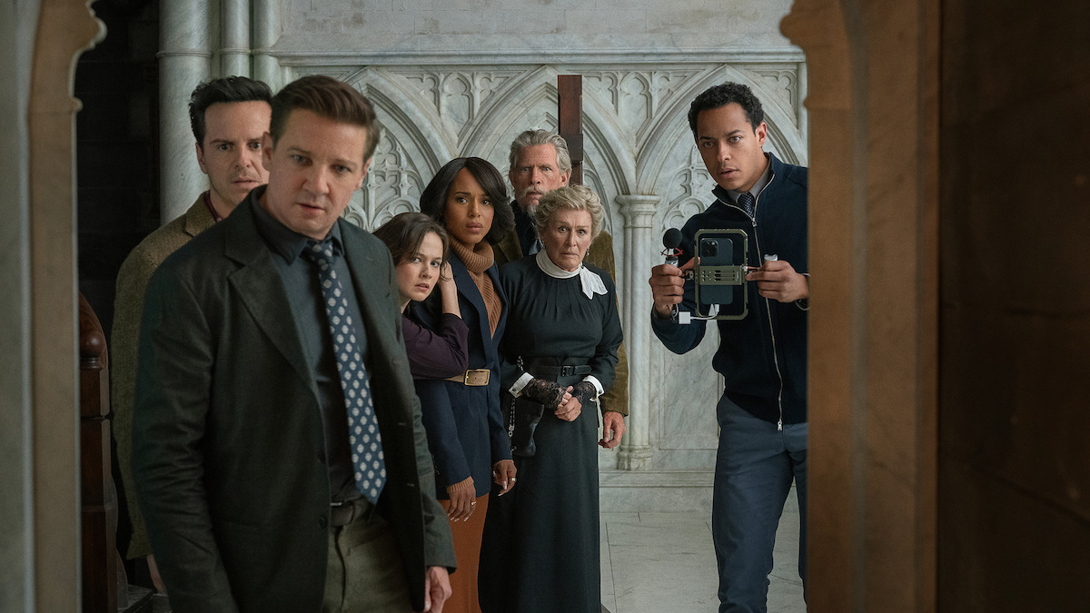 Six people in formal attire stand in a stone hallway with arched carvings, looking intensely at something off-frame; one holds a camera, creating a tense, mysterious mood.