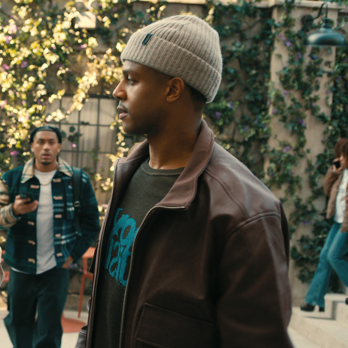 Man in a brown leather jacket and gray beanie walks through an outdoor area with flowering vines on the wall, while people in casual clothing are in the background, one checking his phone.