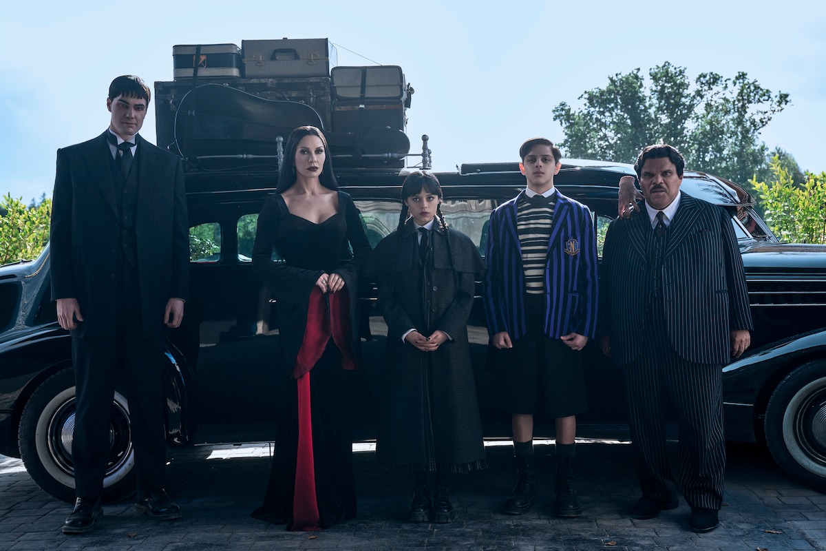 Joonas Suotamo as Lurch, Catherine Zeta-Jones as Morticia Addams, Jenna Ortega as Wednesday, Isaac Ordonez as Pugsley Addams, Thing and Luis Guzmán as Gomez Addams stand in front of a car in ‘Wednesday’ Season 2.