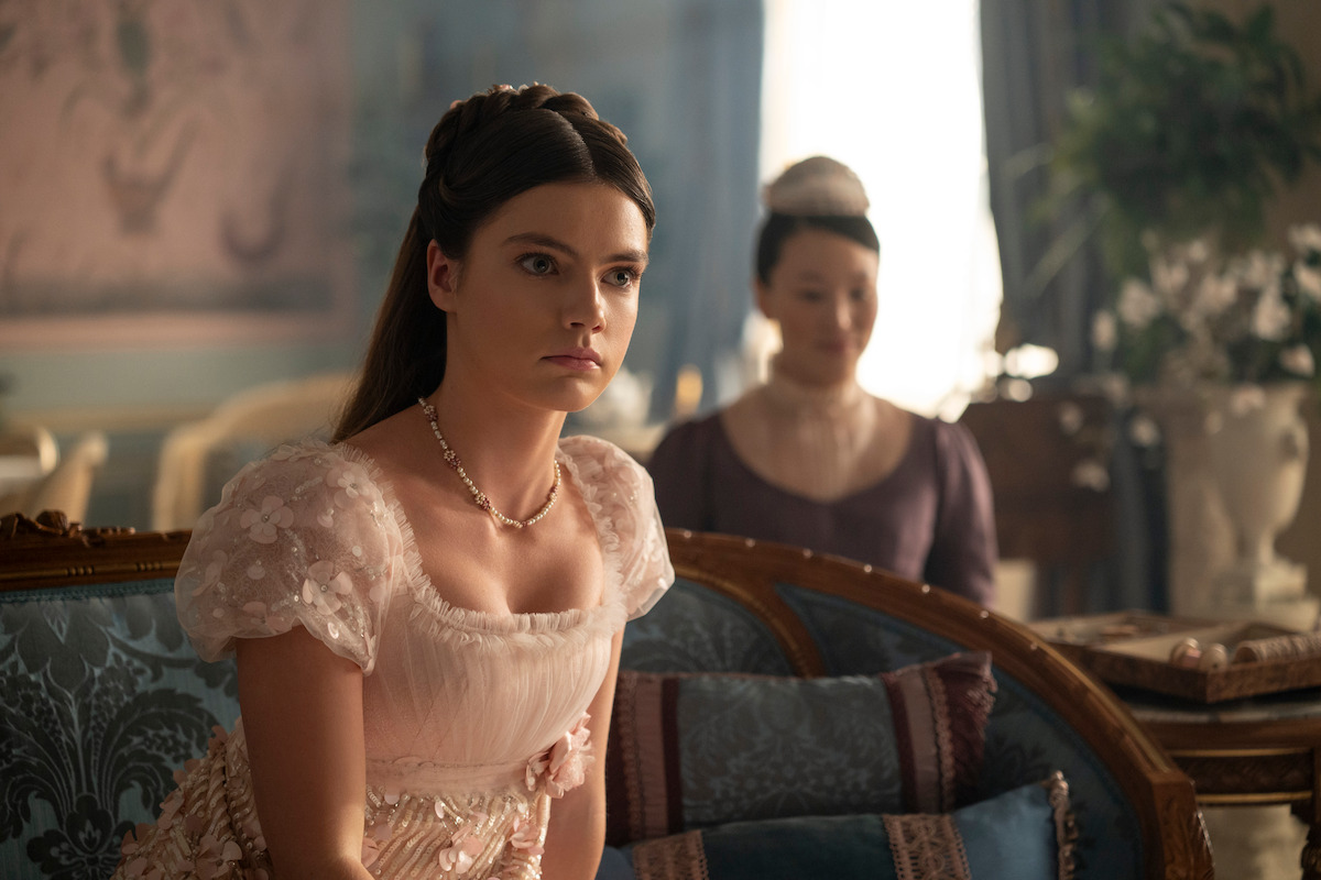 Uma jovem usando um vestido de época rosa claro elegante sentada em um sofá azul estampado, em uma sala com decoração refinada e iluminação natural, e outra mulher ao fundo. O cenário indica um ambiente histórico ou aristocrático.