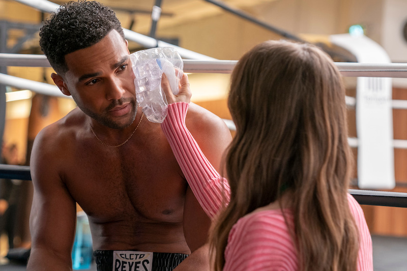 Lucien Laviscount, como Alfie, sentado em um ringue de boxe enquanto Lily Collins, como Emily, segura uma bolsa de gelo contra o seu rosto na temporada 4 de ‘Emily em Paris’