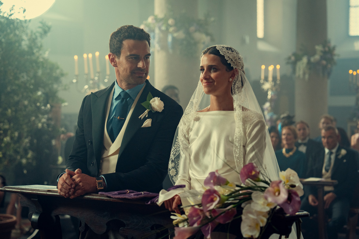 A bride in a white dress and veil sits beside a groom in a suit at an altar, holding a bouquet of flowers in a decorated church filled with guests and candles during a wedding ceremony.