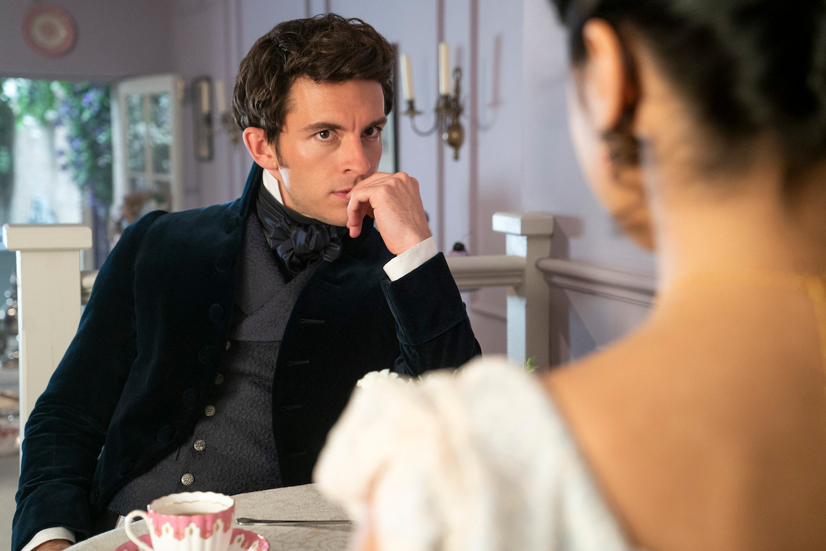 A man in period clothing sits at a table in a softly lit, elegant room, thoughtfully looking at a woman in the foreground. Teacups and pastel decor suggest a historical or Regency-era setting.