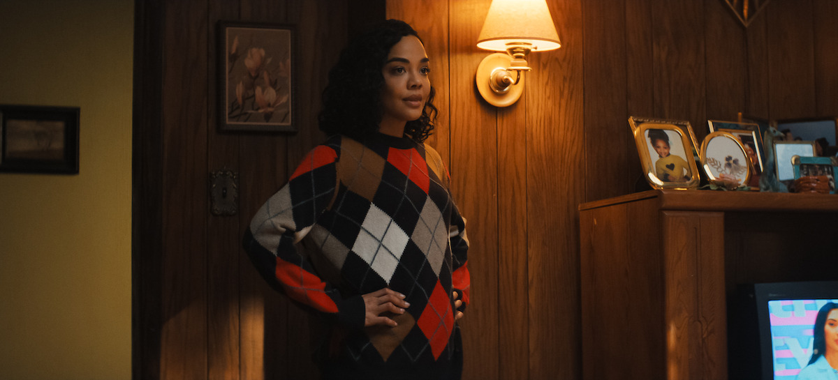 Woman in an argyle sweater stands in a warmly lit, wood-paneled room with family photos and a TV.