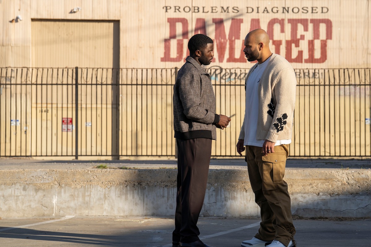 Two men stand facing each other in conversation outdoors, next to a metal fence and concrete curb, with a large building sign in the background reading “PROBLEMS DIAGNOSED DAMAGED” in bold letters.