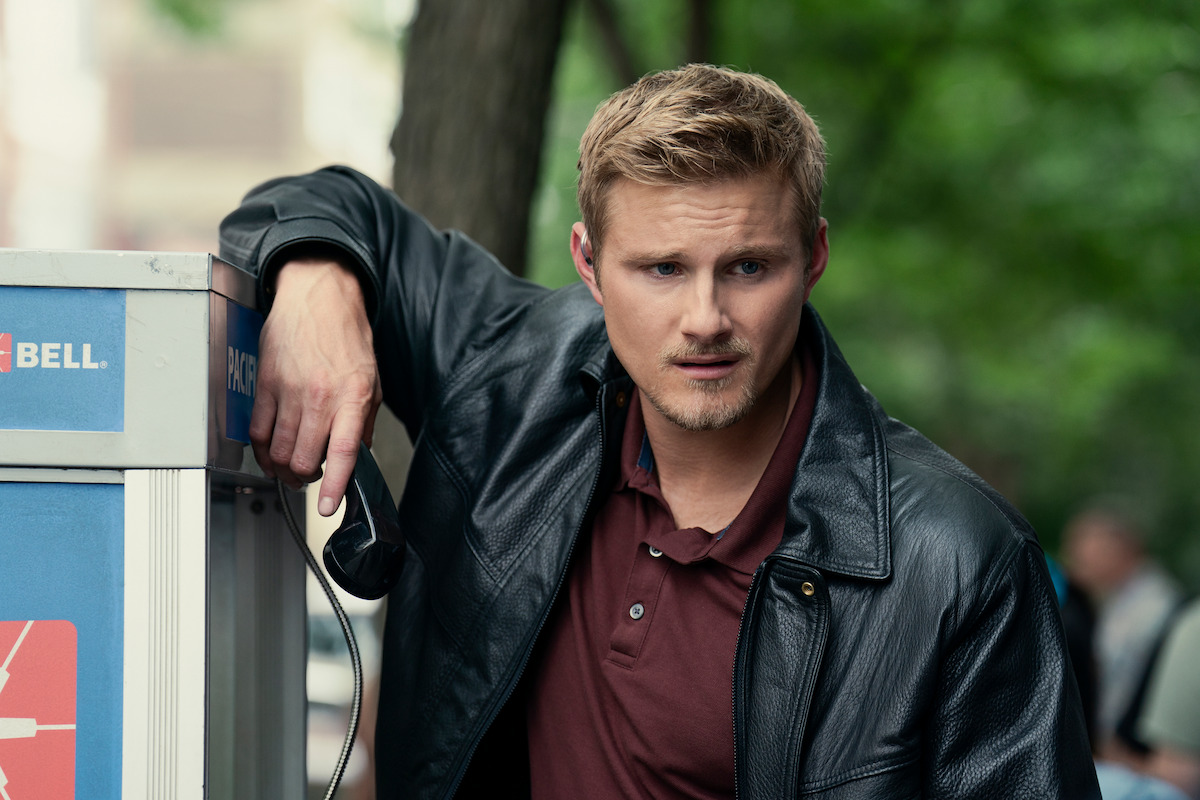Man in leather jacket standing outdoors by a payphone, holding the phone receiver, with trees and blurred people in the background, appearing thoughtful or concerned.