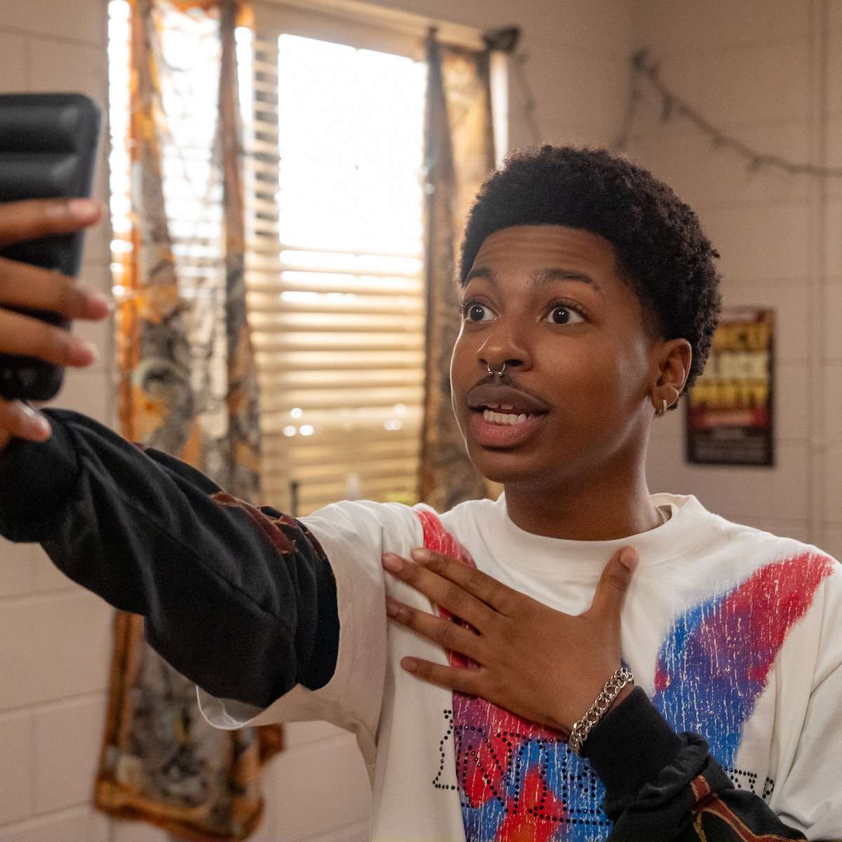 Young person taking a selfie with a surprised facial expression in a sunlit room, hand on chest, decorative curtains and poster in the background.
