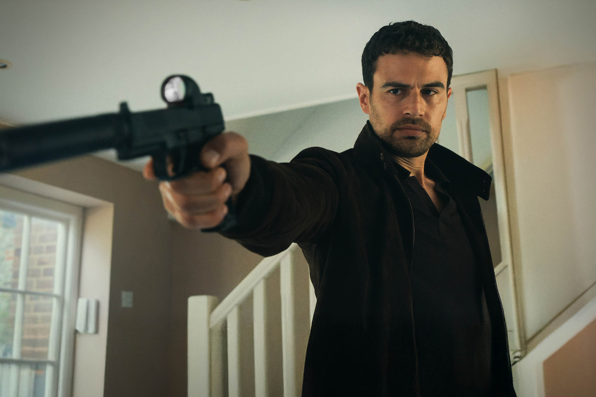 Man indoors in front of a staircase aiming a silenced handgun with a serious expression, wearing a dark jacket, with natural light coming from a window on the left.