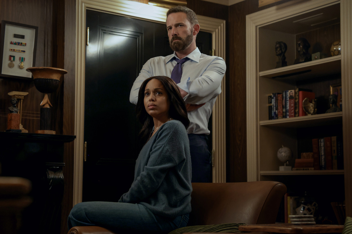 A woman sits on a leather chair looking concerned, while a man stands behind her with arms crossed in a dimly lit office or library filled with shelves, books, and decorative objects.