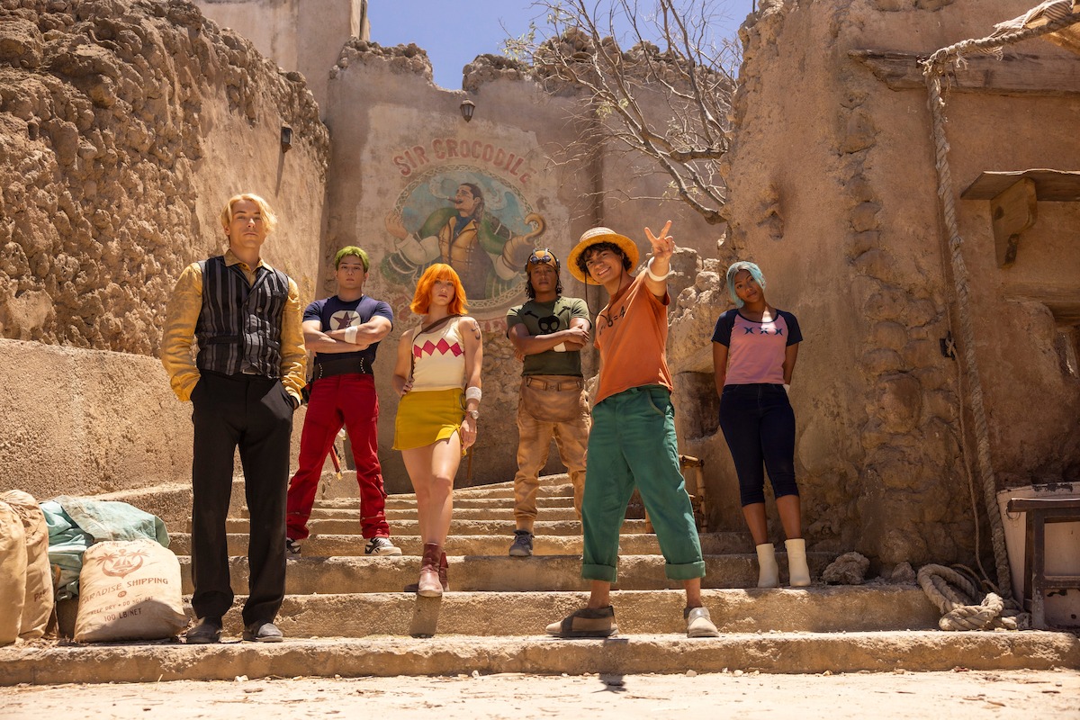 Six people in colorful, varied costumes stand on stone steps in front of a weathered building with a mural, under bright sunlight in a rustic, old village setting.