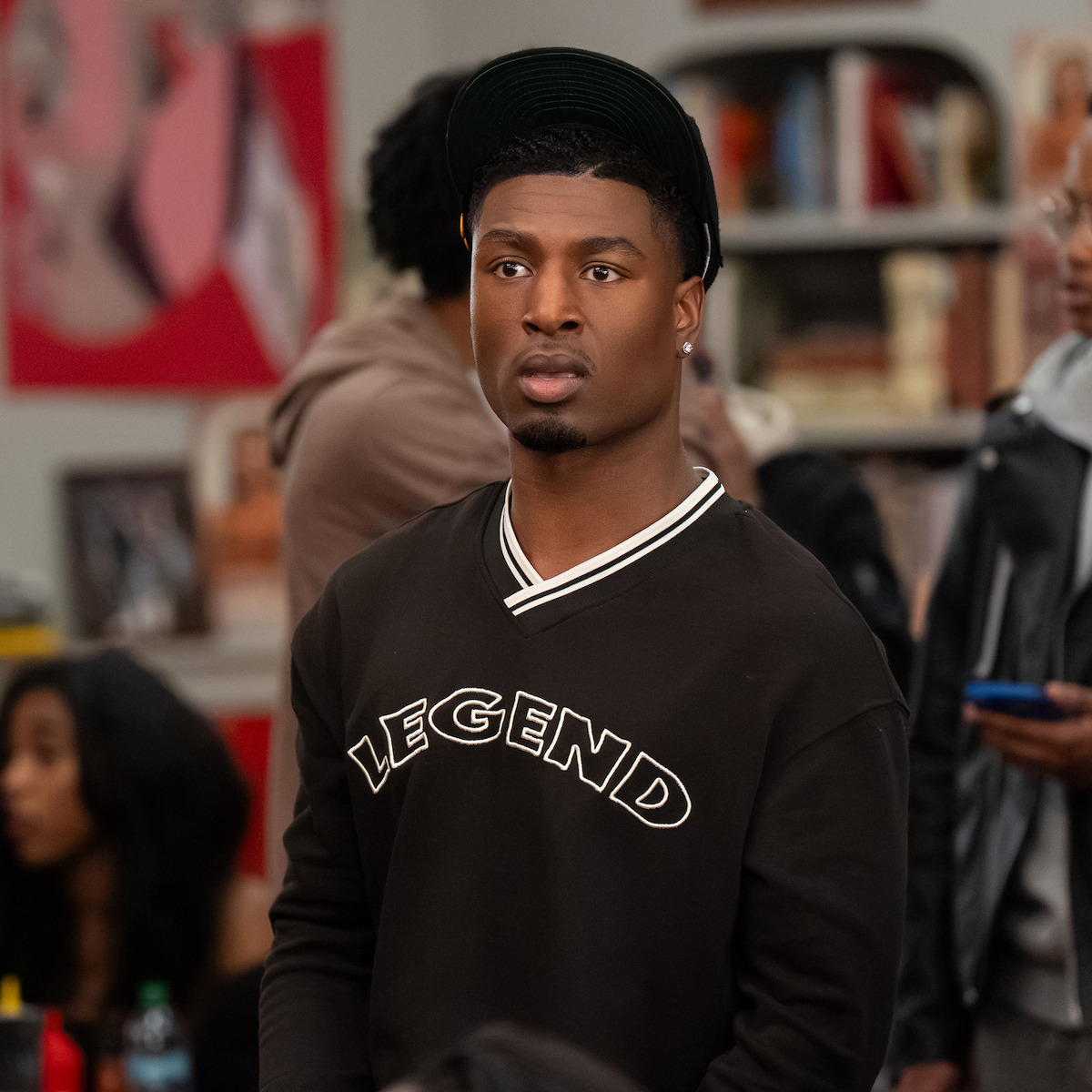 A young man wearing a black sweatshirt with "LEGEND" text stands indoors, surrounded by other people in a casual social setting, with bookshelves and abstract art visible in the background.