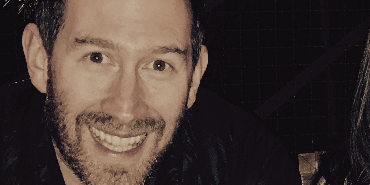 Smiling man with short hair and beard in a dark indoor setting, with a grid-patterned wall in the background.
