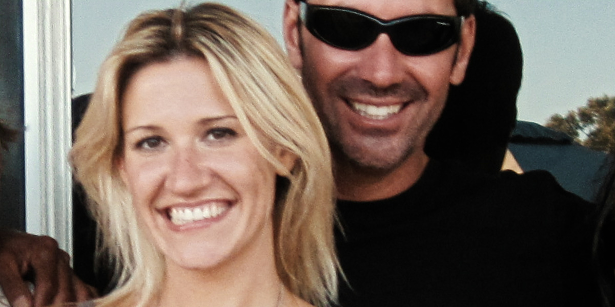 Smiling woman with blonde hair and a man in sunglasses standing close together outdoors, both appearing happy with sunlight and greenery in the background.