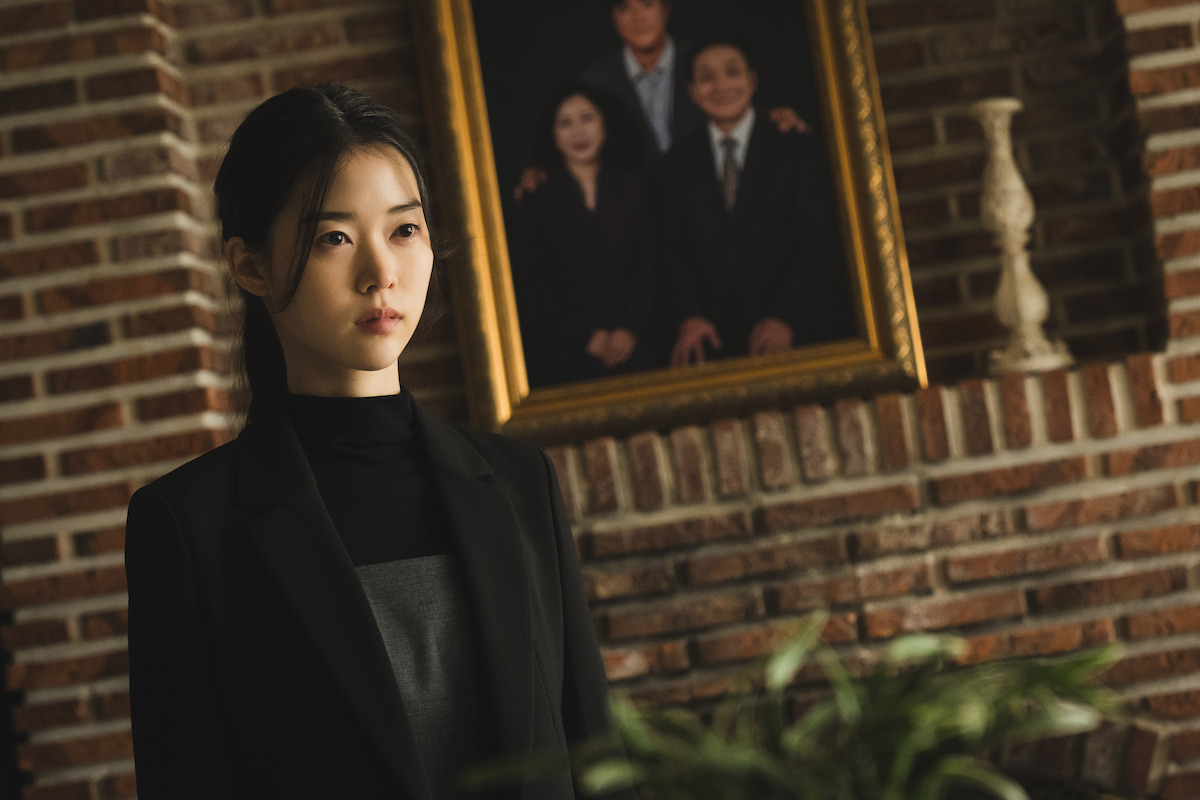 A woman in a black outfit stands indoors near a brick wall with a framed family portrait and a decorative vase, with a green plant in the foreground, creating a somber and elegant setting.