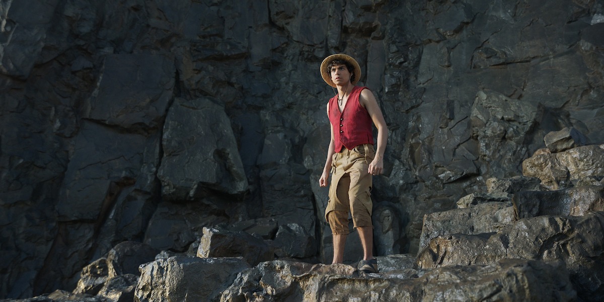 A young person wearing a straw hat, red vest, and rolled-up pants stands on rocky terrain with tall, dark stone cliffs in the background, looking outward in a rugged outdoor environment.