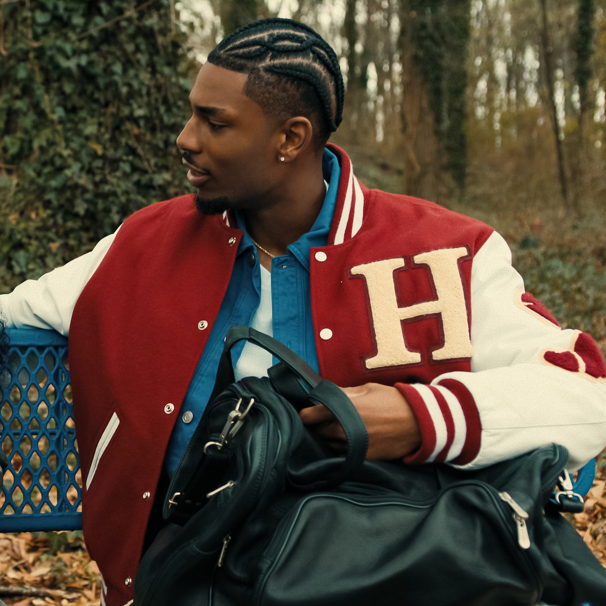 Man wearing a red varsity jacket with an "H," sitting on a blue bench in a wooded park area with fallen leaves, holding a black bag and looking to the side.