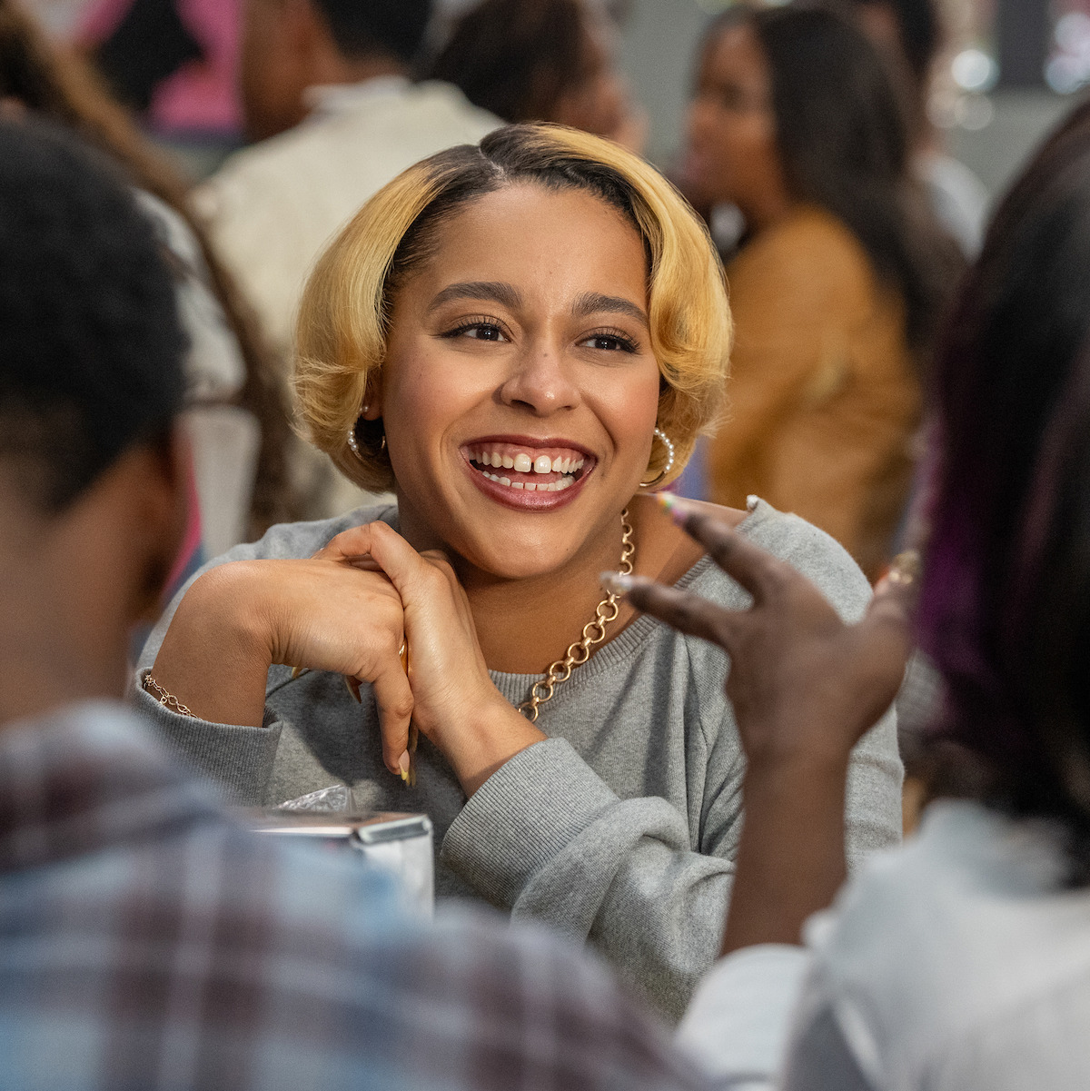 Smiling woman with blonde hair sits at a table in a lively, indoor social setting, engaged in conversation with others around her.