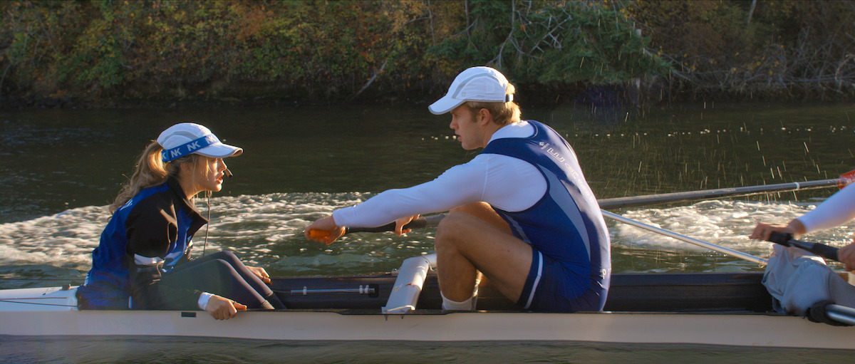 Rowers in blue and white uniforms compete on a calm river, with a female coxswain guiding the boat and trees in the background, sunlight reflecting off the water.