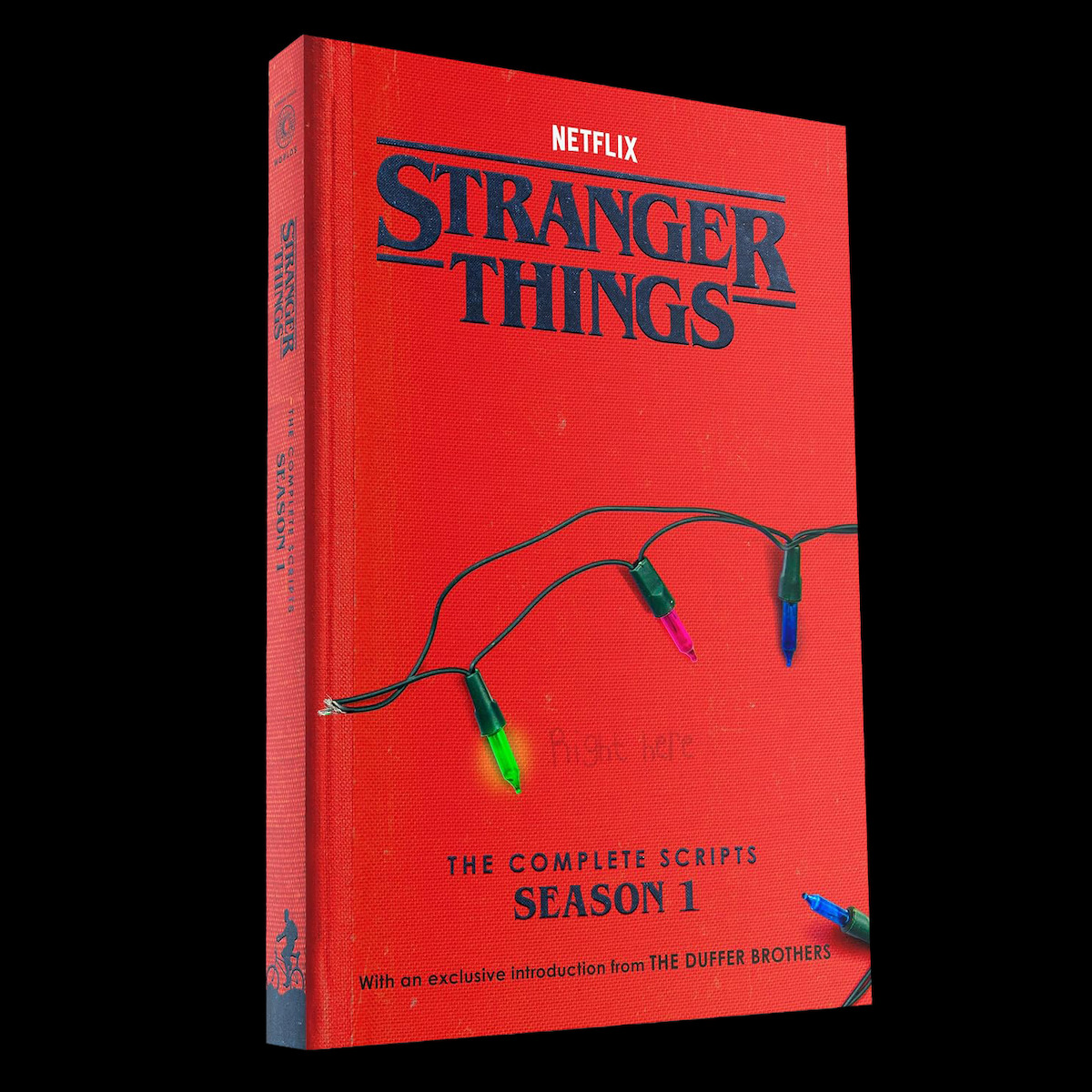 Livro vermelho com o título “Stranger Things: The Complete Scripts Season 1” da Netflix, com luzes de Natal na capa e à frente de um fundo preto. Ambientação misteriosa e nostálgica fazendo referência aos temas da série.