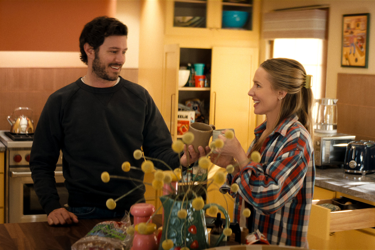 A man and woman smiling and chatting while holding mugs in a cozy, warmly lit kitchen with wooden cabinets, decorative plants, bread, and appliances visible on the counter and shelves.