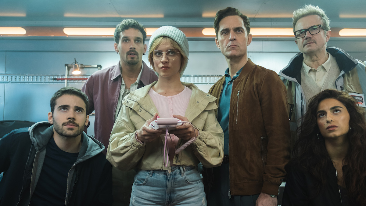 Five people in casual clothing stand and sit closely together indoors, focused intently on something ahead, with a woman in a beanie and glasses holding a device; industrial lighting and shelves in the background.